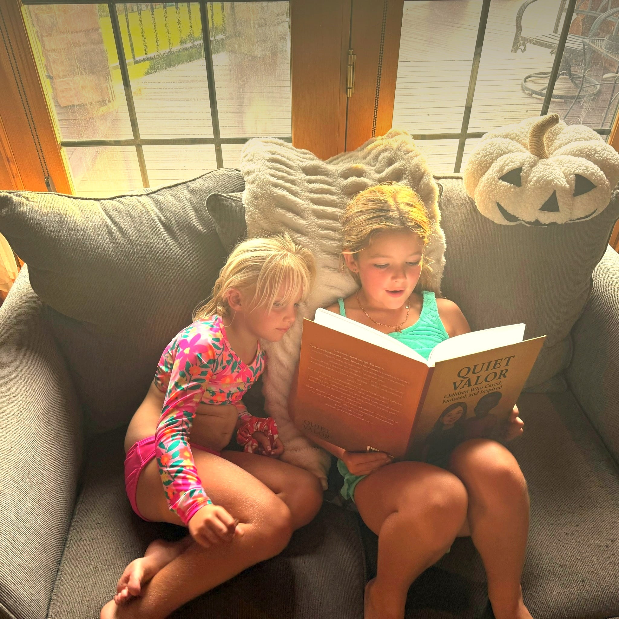 Two young girls sit on a couch, engrossed in a book titled "Quiet Valor," with sunlight streaming through nearby windows, creating a cozy reading atmosphere.
